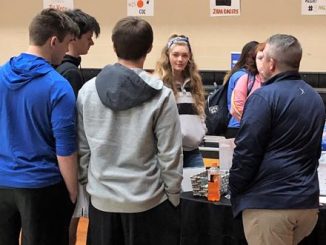 Students are shown surrounding an exhibitors booth at the Lynn Camp Career Fair asking questions and looking at items on display.